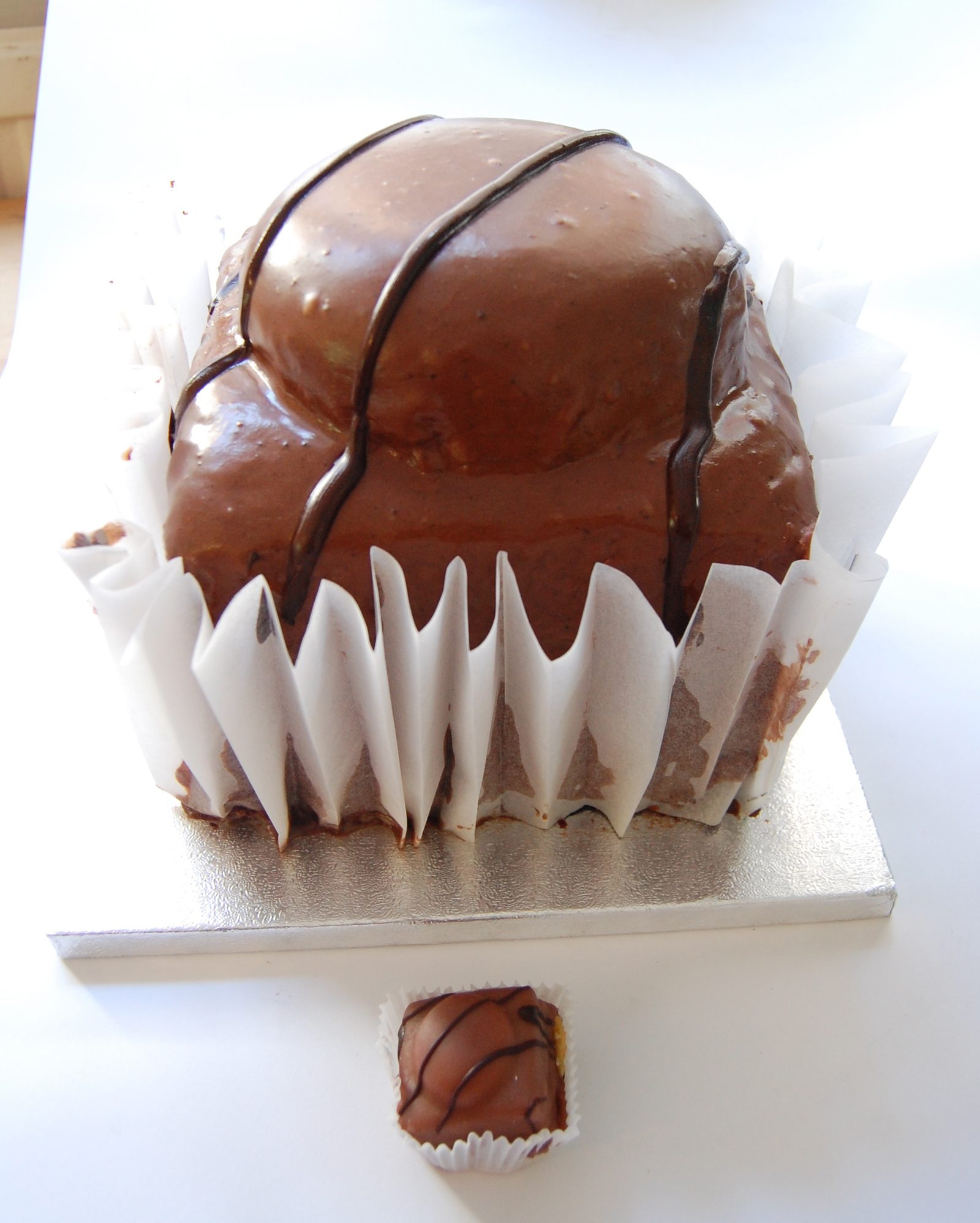 Giant chocolate-glazed fondant fancy in a paper case on a cake board, a normal one in front
